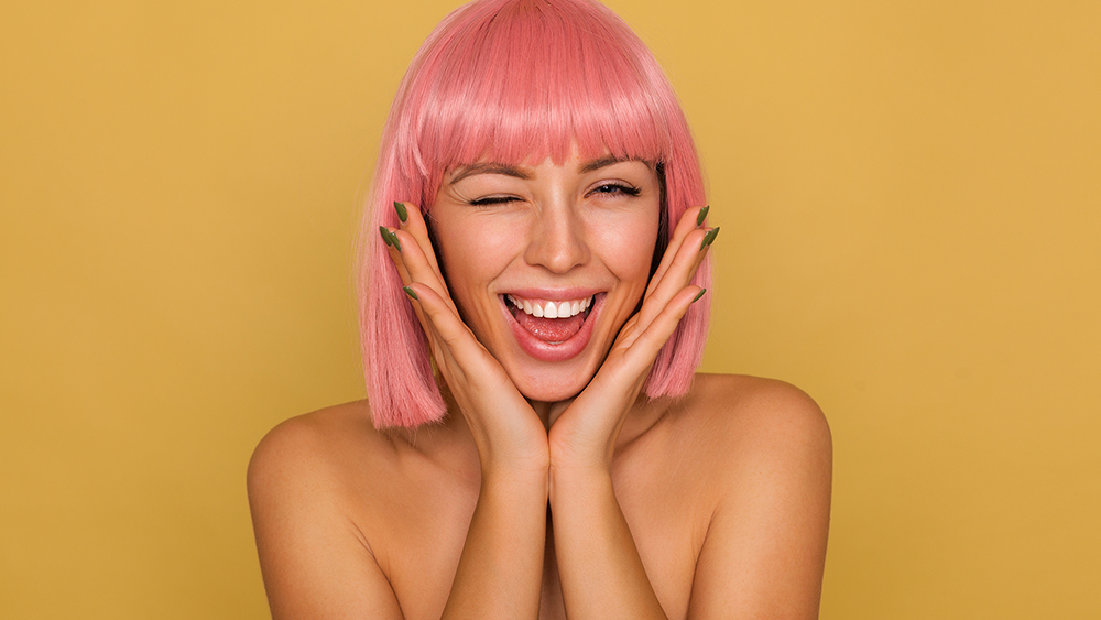 Indoor shot of young attractive pink haired woman with bob hairstyle giving wink at camera and smiling happily while leaning chin on raised palms, isolated over mustard background
